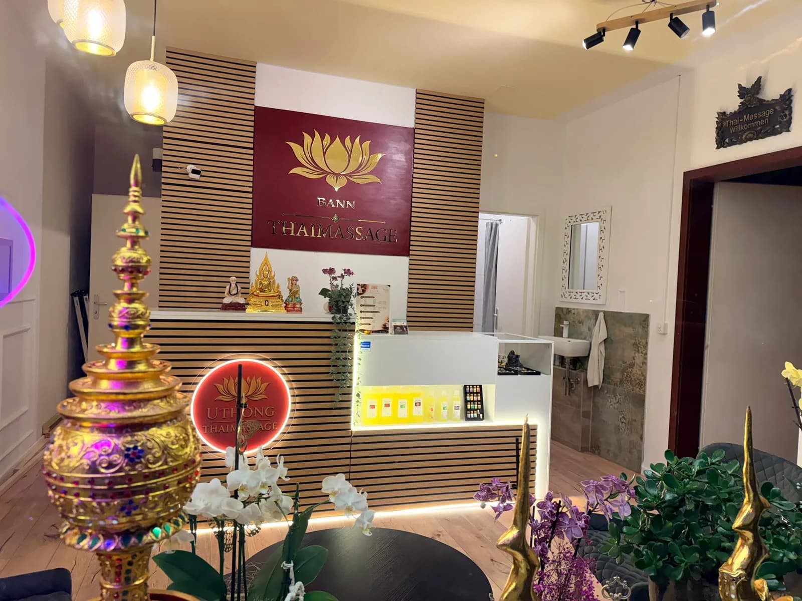 Empfang von BANN Thaimassage in Basel Empfang von BANN Thaimassage in Basel: Tresen aus Holzlamellen mit goldenem Lotus-Logo, davor Orchideen und goldene thailändische Ziergefässe