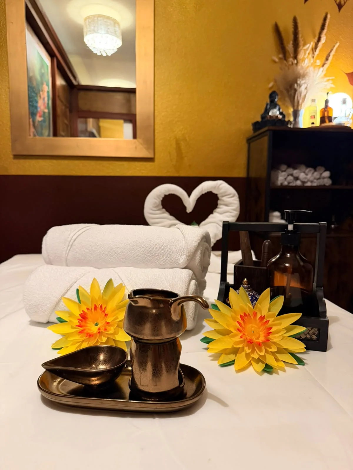 Vorbereitete Massageliege mit Handtüchern und Lotusblüten Vorbereitete Massageliege bei BANN Thaimassage in Basel: gerollte Handtücher, gelbe Lotusblüten, ein bronzefarbenes Kännchen und ein Herz aus Handtüchern