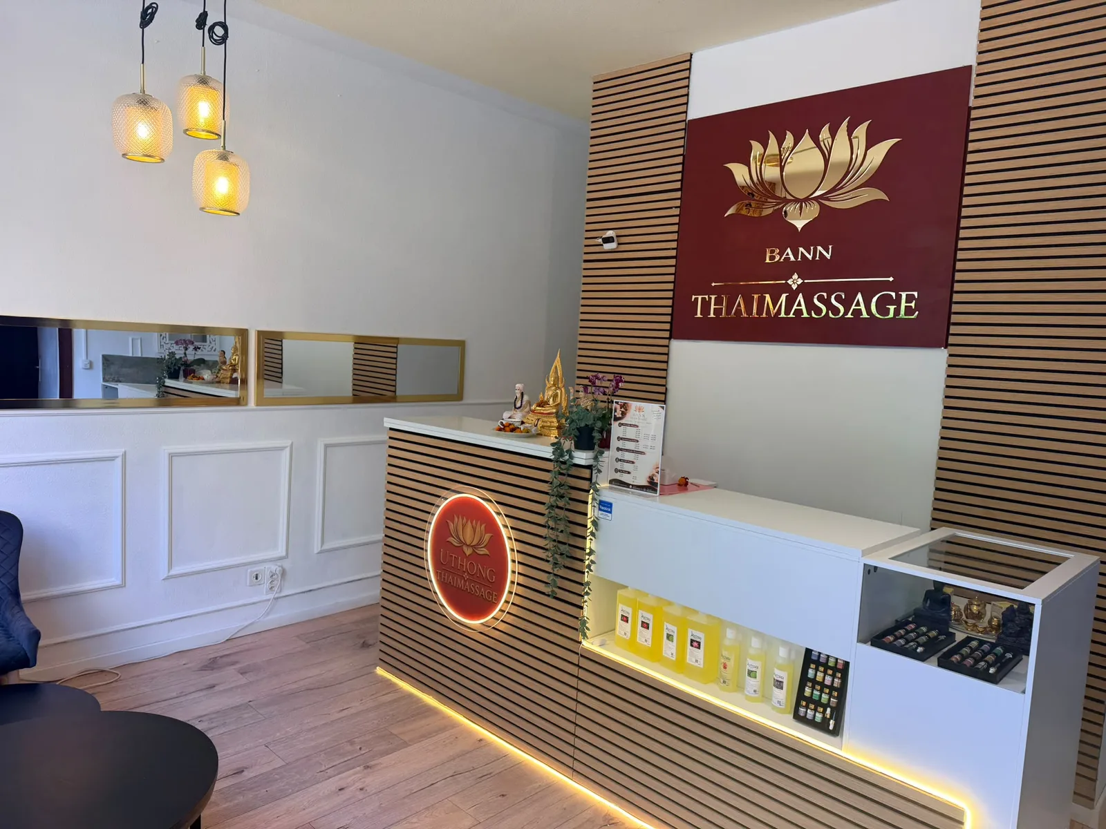 Empfangsbereich von BANN Thaimassage in Basel Empfangsbereich von BANN Thaimassage in Basel mit Tresen aus Holzlamellen, goldenem Lotus-Logo auf rotem Grund und Pendelleuchten